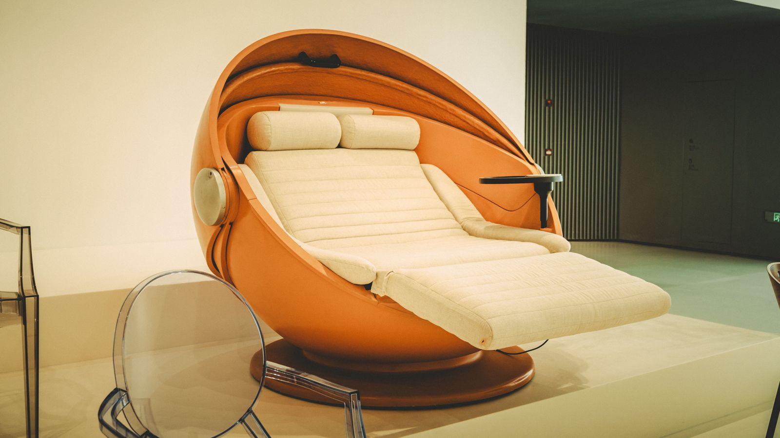 Orange spherical lounge pod with a cream quilted reclining seat, bolster cushions and side tray on display